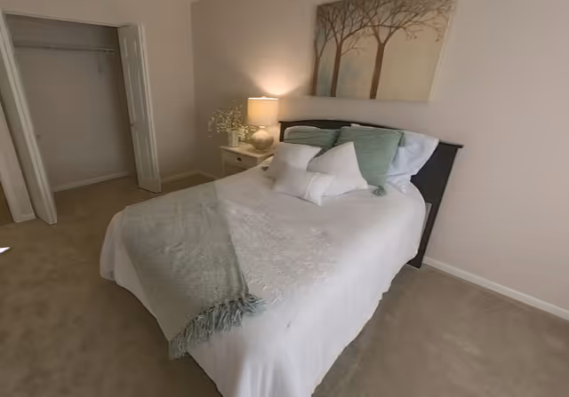 Neatly made bed with pillows and a throw in a softly lit bedroom with a nightstand, lamp, wall art, and an open closet.