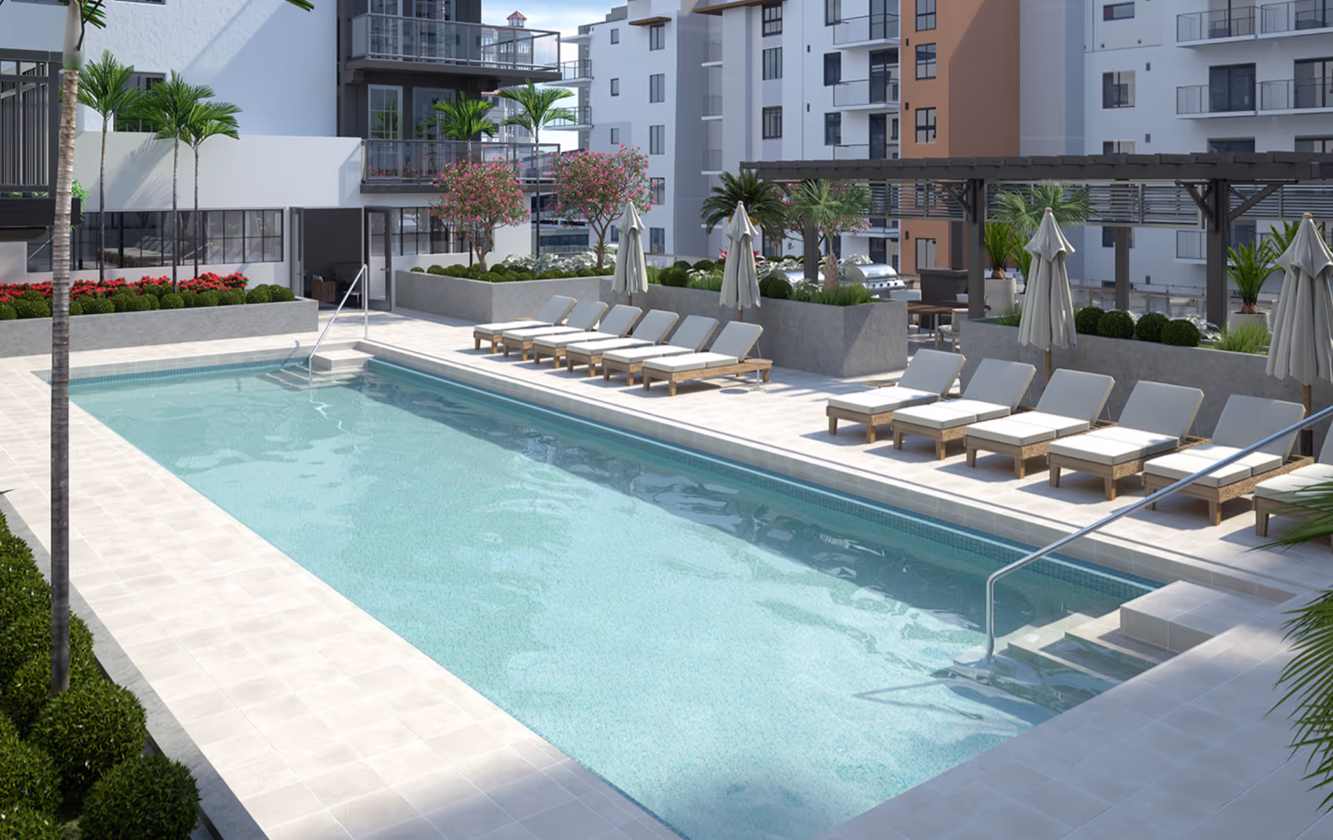 Outdoor swimming pool area with lounge chairs and umbrellas arranged along the poolside. The pool is surrounded by tiled flooring and landscaped with palm trees, bushes, and flowering plants. Modern multi-story buildings are visible in the background.