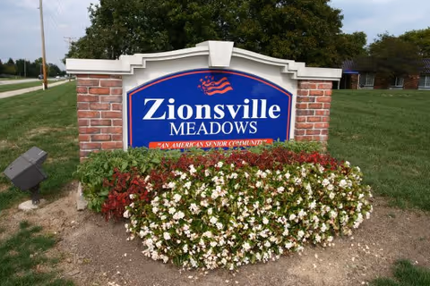 A large blue sign with white text reading 'Zionsville Meadows' and a smaller red banner below stating 'An American Senior Community', set in a brick frame with flowers planted in front and a grassy area with trees in the background.