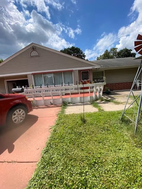 Exterior view of a single-story residential building with beige siding and a red brick lower section. There is a white railing along a small porch area, a red car parked on a driveway, and a grassy lawn in the foreground. The sky is partly cloudy with blue patches visible.