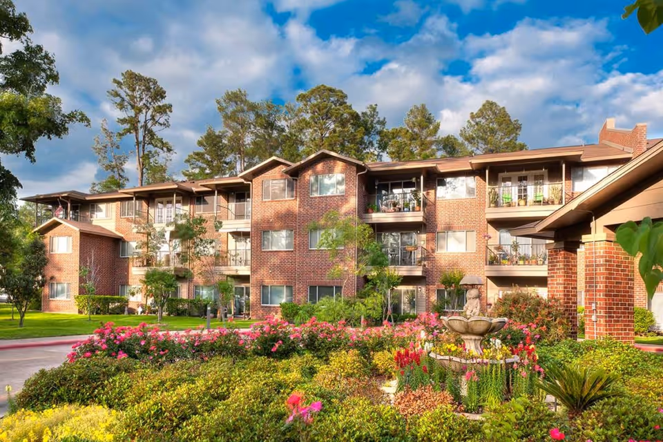 Exterior view of a multi-story brick retirement community building surrounded by well-maintained gardens with colorful flowers, shrubs, and a decorative fountain in the foreground under a partly cloudy sky.