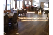 A sunlit room with wooden flooring featuring several tables and chairs arranged for dining. Large windows on one side allow natural light to fill the space, and there are plants and decorative items near the far wall.