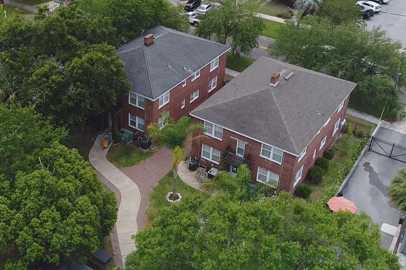 Aerial view of two brick residential buildings surrounded by trees and greenery, with a curved sidewalk and a small patio area with chairs and a table. A gated driveway is visible on the right side.