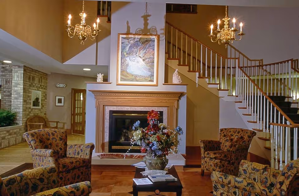 A cozy living room area with patterned armchairs arranged around a coffee table holding a vase of colorful flowers and some magazines. A fireplace with a wooden mantel is centered on the back wall, above which hangs a framed painting. Two ornate chandeliers hang from the ceiling, and a staircase with wooden handrails and white balusters ascends to the right.