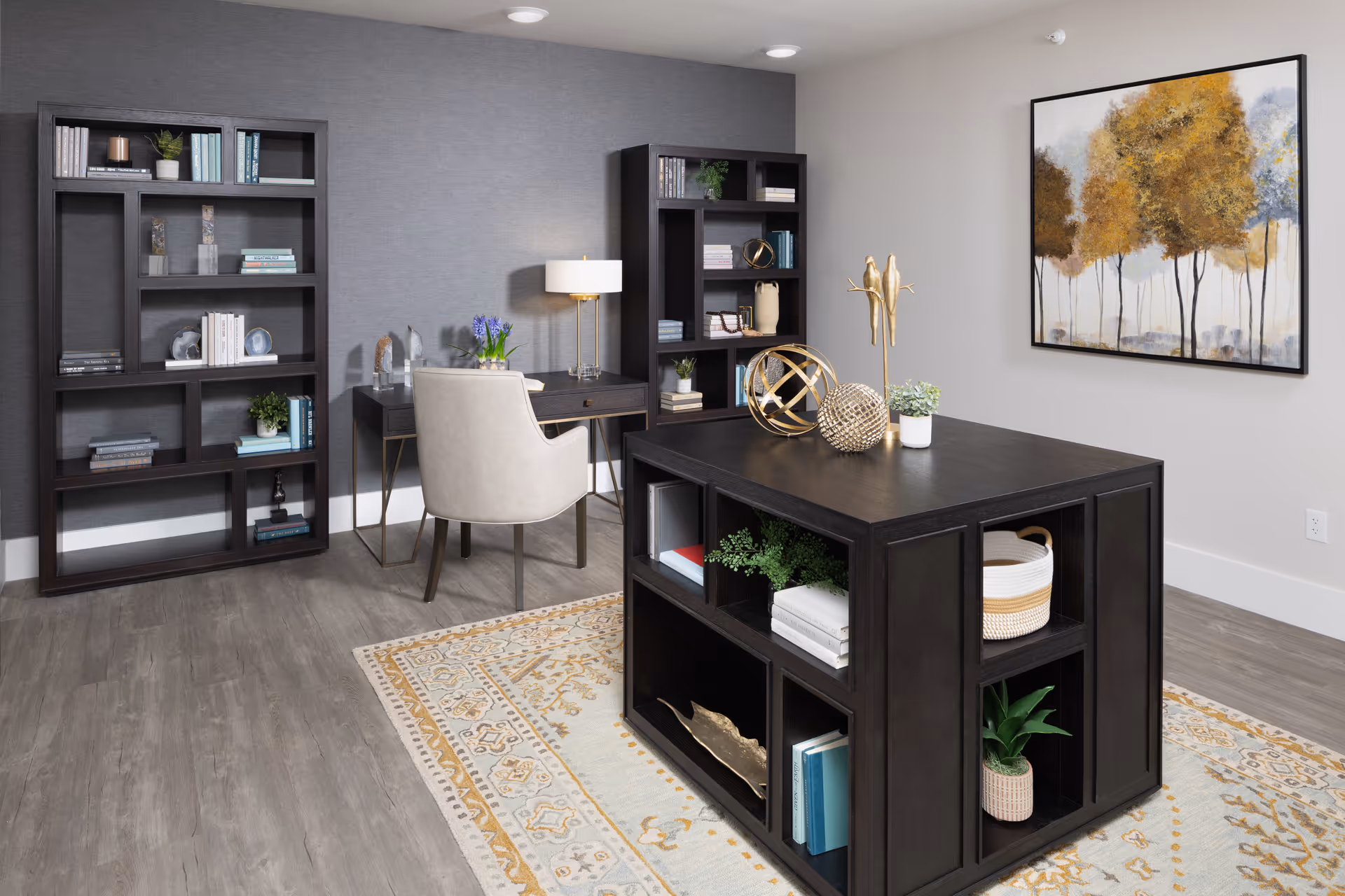 A modern office or study room with two dark wood bookshelves filled with books and decorative items, a desk with a beige upholstered chair and a lamp, and a large dark wood island with open shelving containing plants, books, and decorative objects. The room has light gray walls, a patterned area rug, and a framed painting of trees with yellow and brown foliage on the wall.