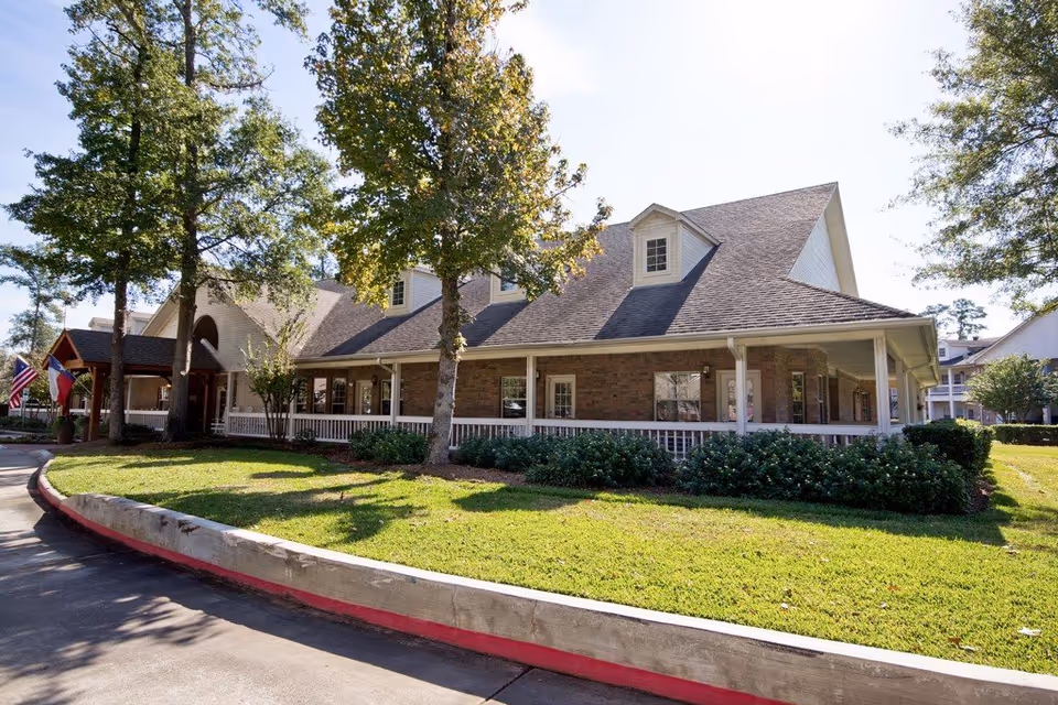 Exterior view of a single-story brick building with a large sloped roof and dormer windows, surrounded by green grass, bushes, and tall trees under a clear sky. There is a covered porch with white railings and a driveway with a red curb in front.