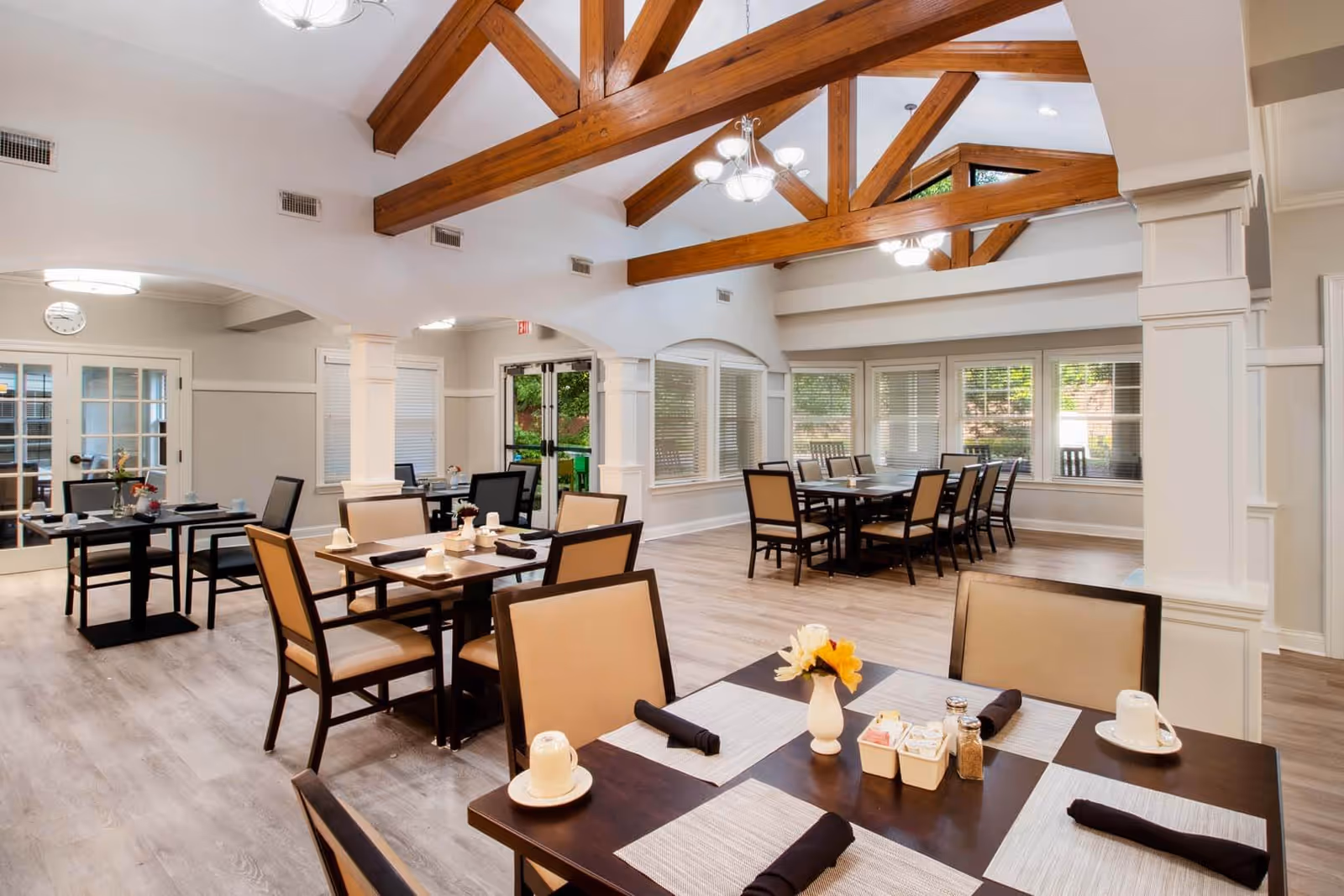 A bright and spacious dining room with wooden beams on the ceiling, multiple tables set with cups, napkins, and small flower vases, beige cushioned chairs, large windows with blinds, and light wood flooring.