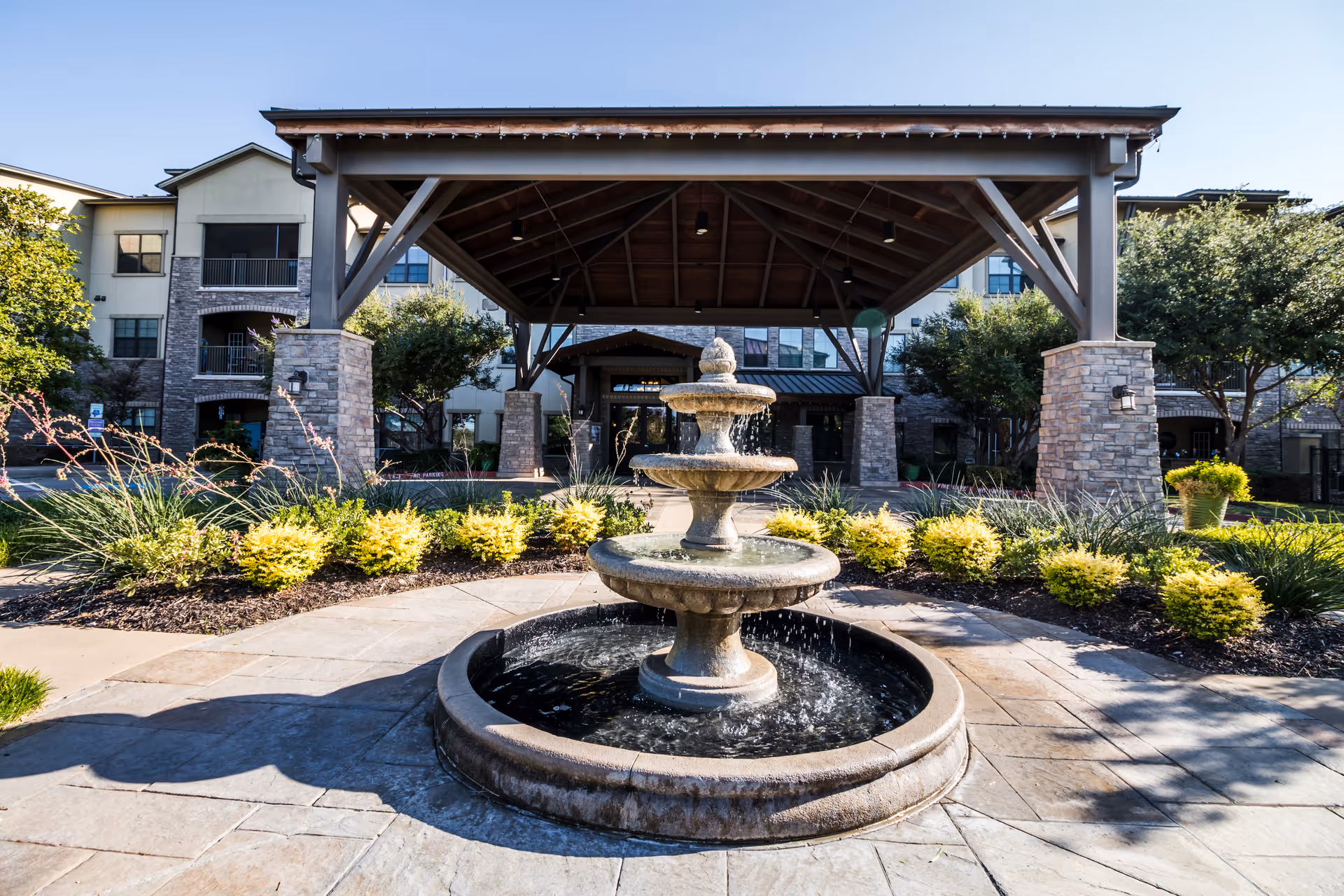 Three-tiered stone fountain in front of a covered porte-cochere entrance to a multi-story senior living building with landscaping.