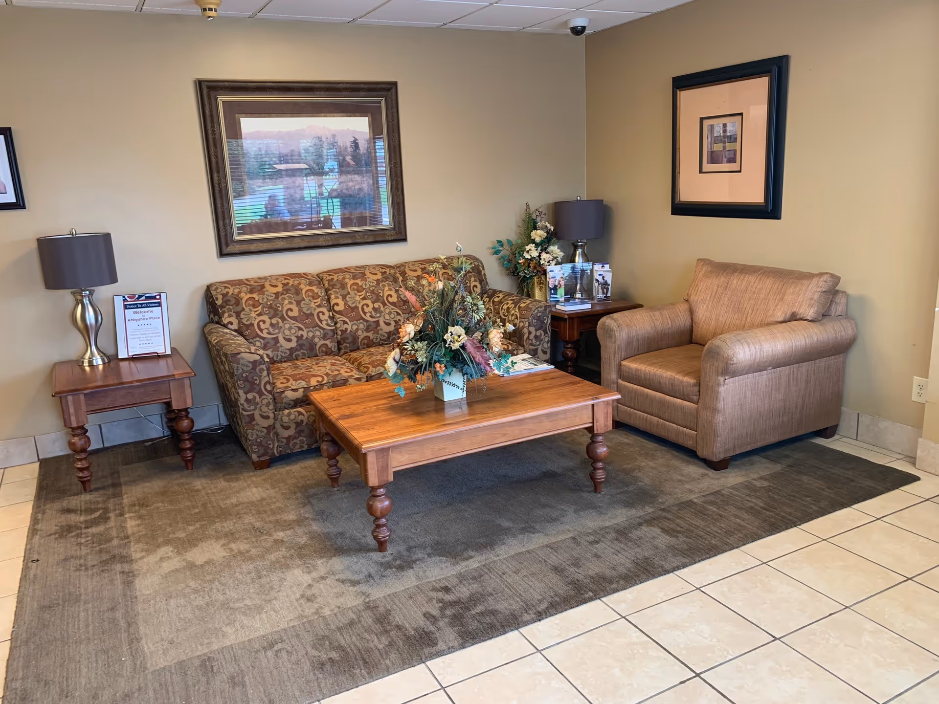 A cozy seating area with a patterned sofa, a beige armchair, and a wooden coffee table with a floral arrangement on top. There are two side tables with lamps and framed artwork on the beige walls. The floor is tiled with a large area rug under the furniture.