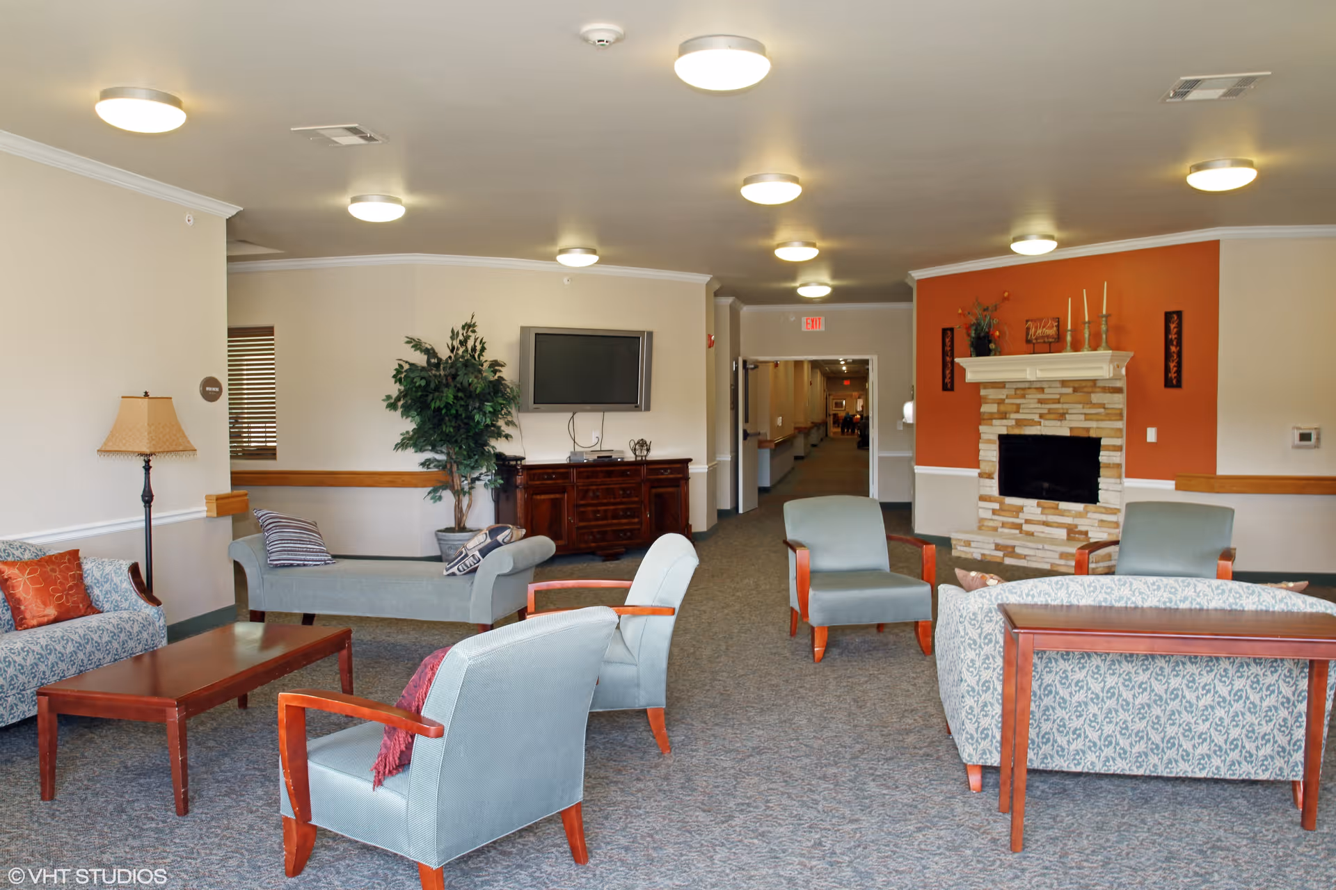 A spacious senior living facility common area with multiple upholstered chairs and sofas arranged around a wooden coffee table. There is a stone fireplace with decorative items on the mantel against an orange accent wall. A flat-screen TV is mounted on the wall above a wooden cabinet, and a potted plant is placed nearby. The room is well-lit with ceiling lights and has a carpeted floor. A hallway is visible in the background.
