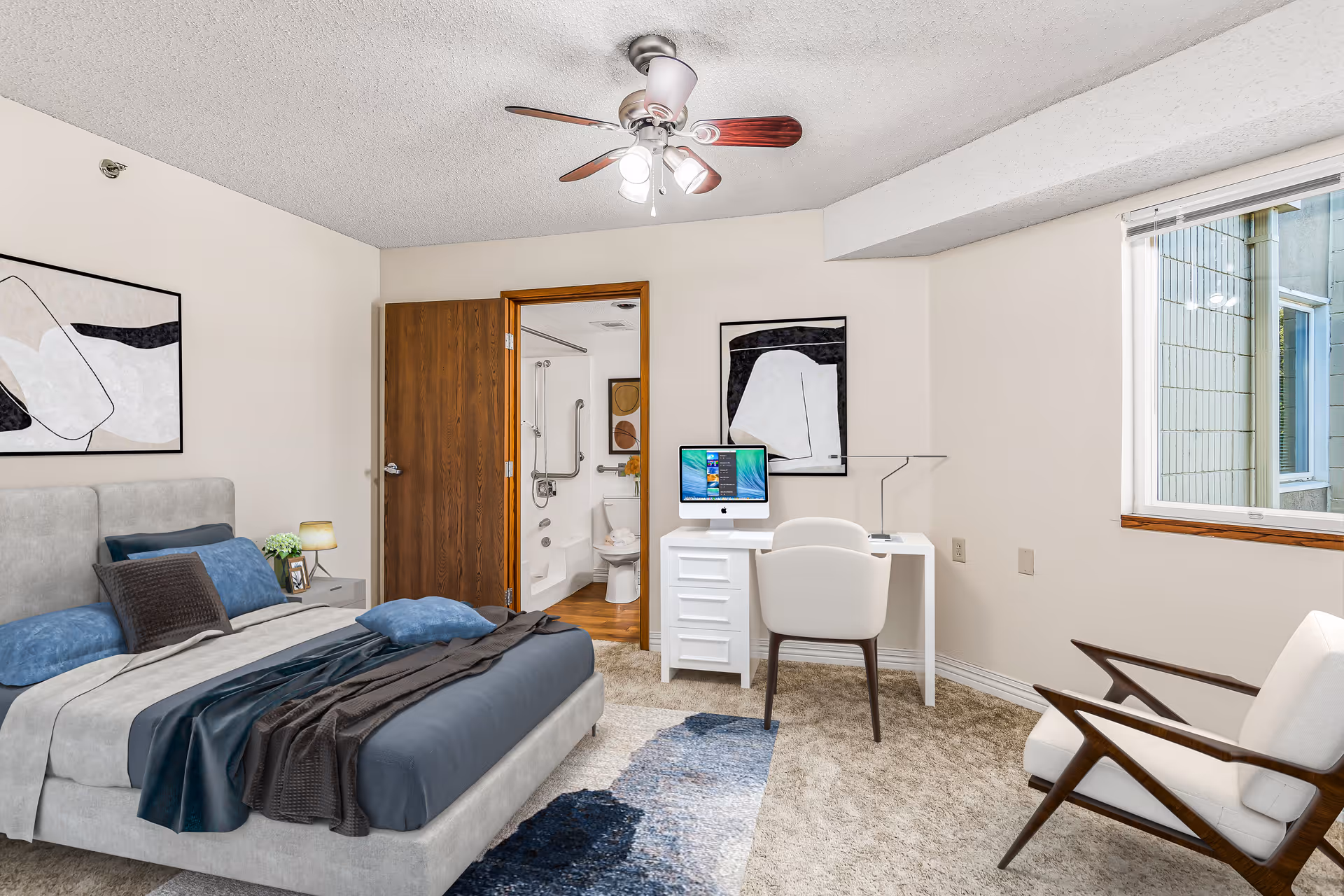 A bedroom in Lawrence Presbyterian Manor featuring a bed with blue and gray bedding, a nightstand with a lamp and plant, a white desk with a computer and chair, an armchair, a ceiling fan with lights, and an open door leading to a bathroom with grab bars and a toilet.