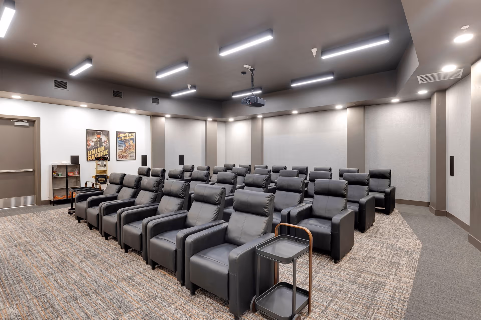 A theater-style media room with rows of black reclining chairs, a ceiling-mounted projector, and movie posters on the wall.