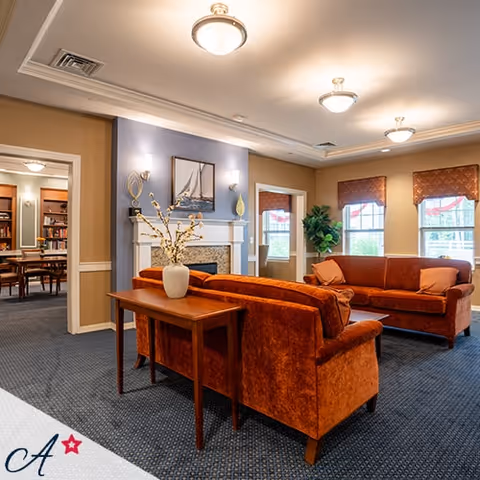 A cozy living room area with two rust-colored sofas facing each other, a wooden console table with a white vase holding decorative branches, a fireplace with a sailboat painting above it, and windows with red valances letting in natural light. The room has beige walls, blue carpeting, and ceiling lights. An adjacent room with bookshelves and a table is visible through an open doorway.
