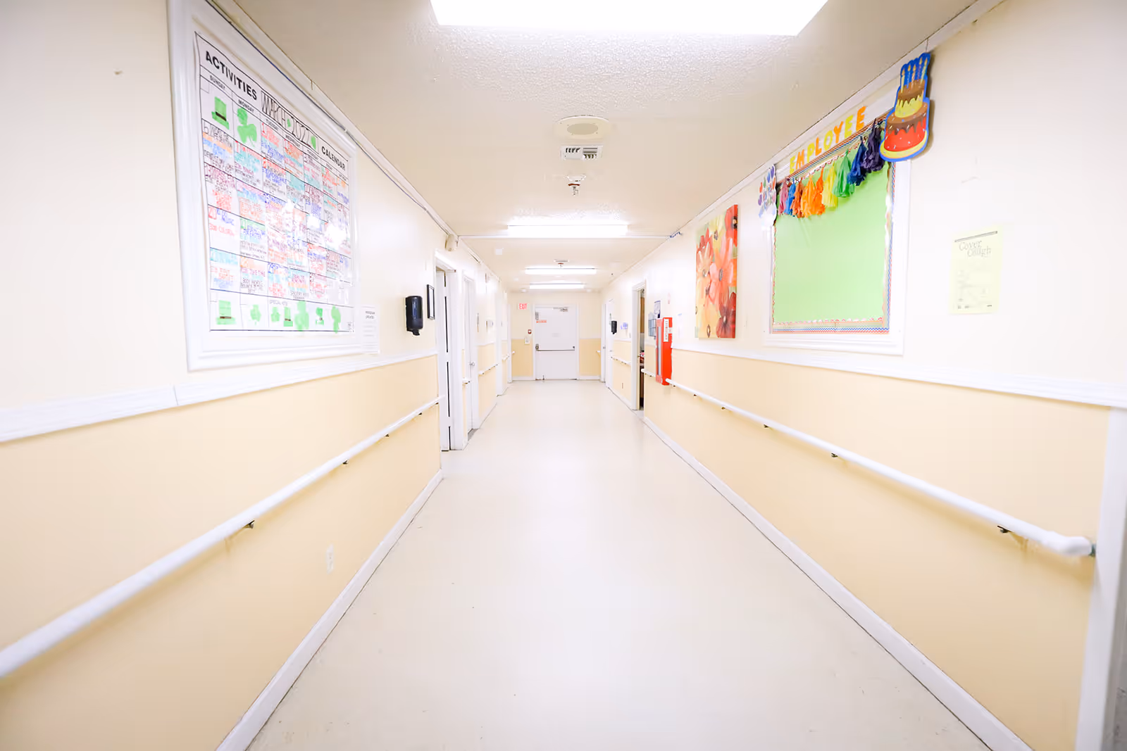 A brightly lit hallway in a rehabilitation and healthcare center with beige and white walls, handrails on both sides, bulletin boards, and an activities calendar displayed on the walls.