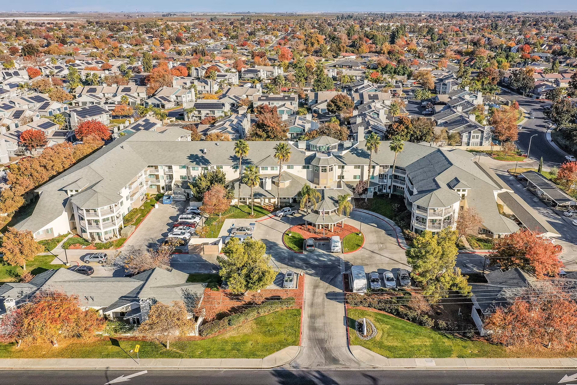 Aerial view of a senior living complex with a circular driveway, parked cars, and surrounding residential neighborhood in autumn.