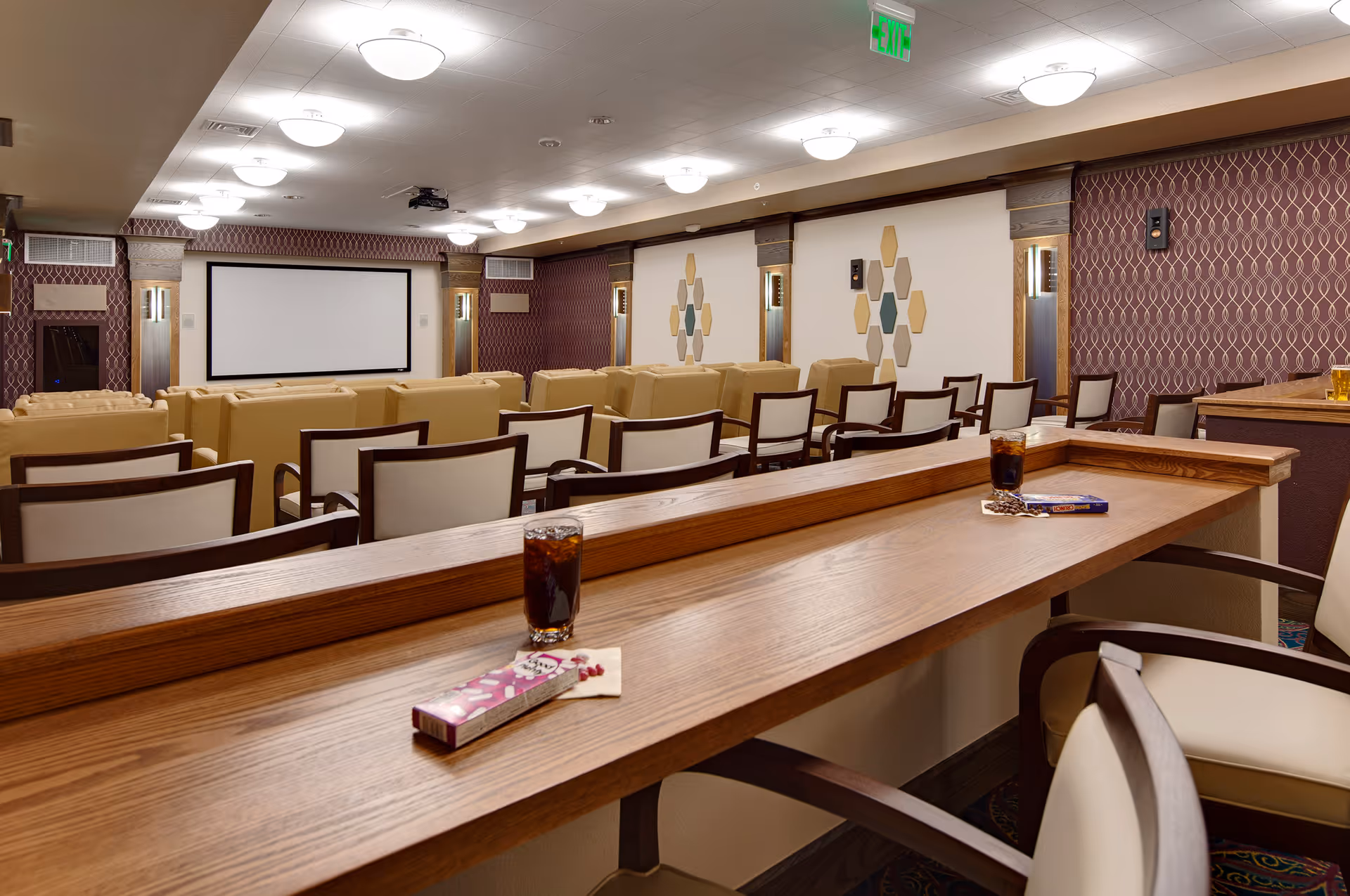 Interior view of a theater-style room with rows of beige cushioned chairs and a large white projection screen at the front. The room has patterned wallpaper, wall-mounted lights, and a wooden counter with chairs in the foreground, where two glasses of dark soda and a candy bar are placed.