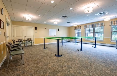 Well-lit interior multipurpose room with a green table tennis setup, chairs along the wall, and windows with patterned valances.