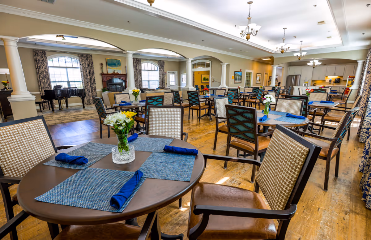 Bright communal dining room with round tables set with blue napkins and flower centerpieces, columns, a piano and an open kitchen in the background.