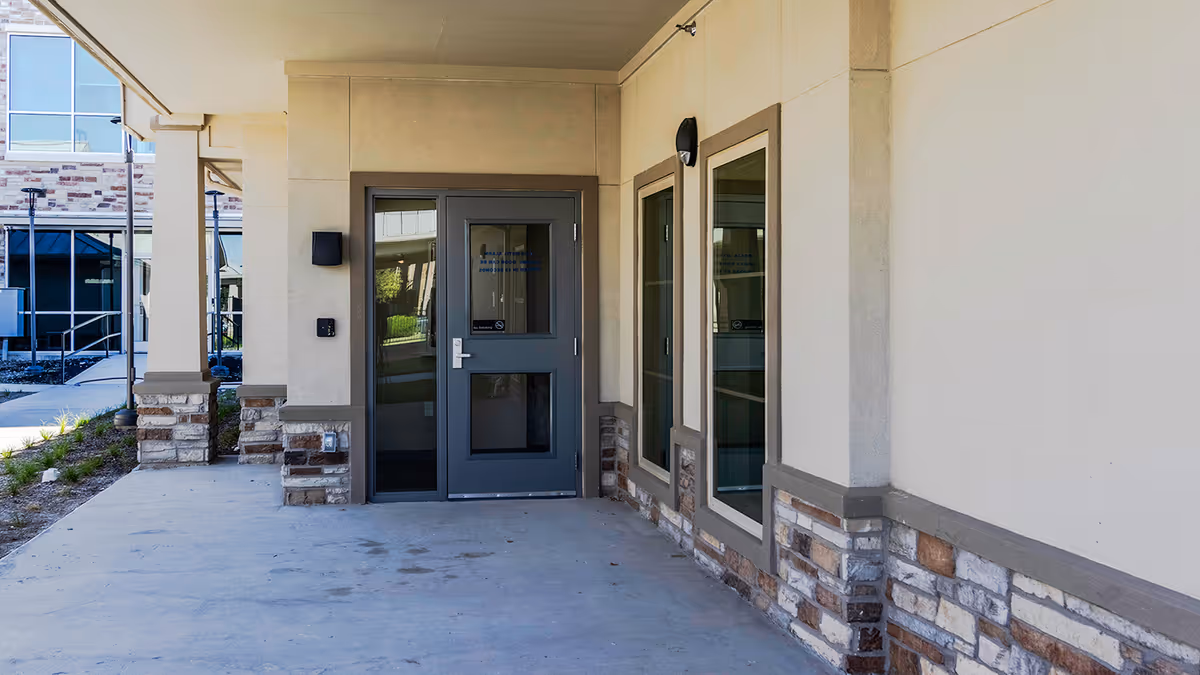 Covered entrance area of a senior living facility with a gray door and adjacent windows, beige walls with stone accents, and outdoor lighting fixtures.