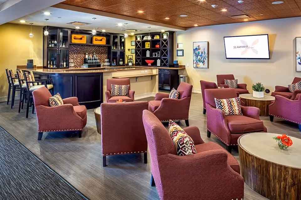 A cozy lounge area with multiple maroon upholstered armchairs arranged around circular wooden tables, each table decorated with small plants or flowers. In the background, there is a bar with high chairs, shelves with decorative items, and a mounted flat-screen TV on the wall displaying the word 'ASPIRED'. The room has warm lighting and a wooden ceiling.