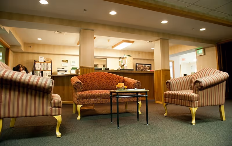 A cozy seating area in a senior living facility with two striped armchairs and a patterned loveseat arranged around a small black coffee table. Behind the seating area is a reception desk with office supplies and a person partially visible. The room has warm lighting and beige walls with wood paneling.