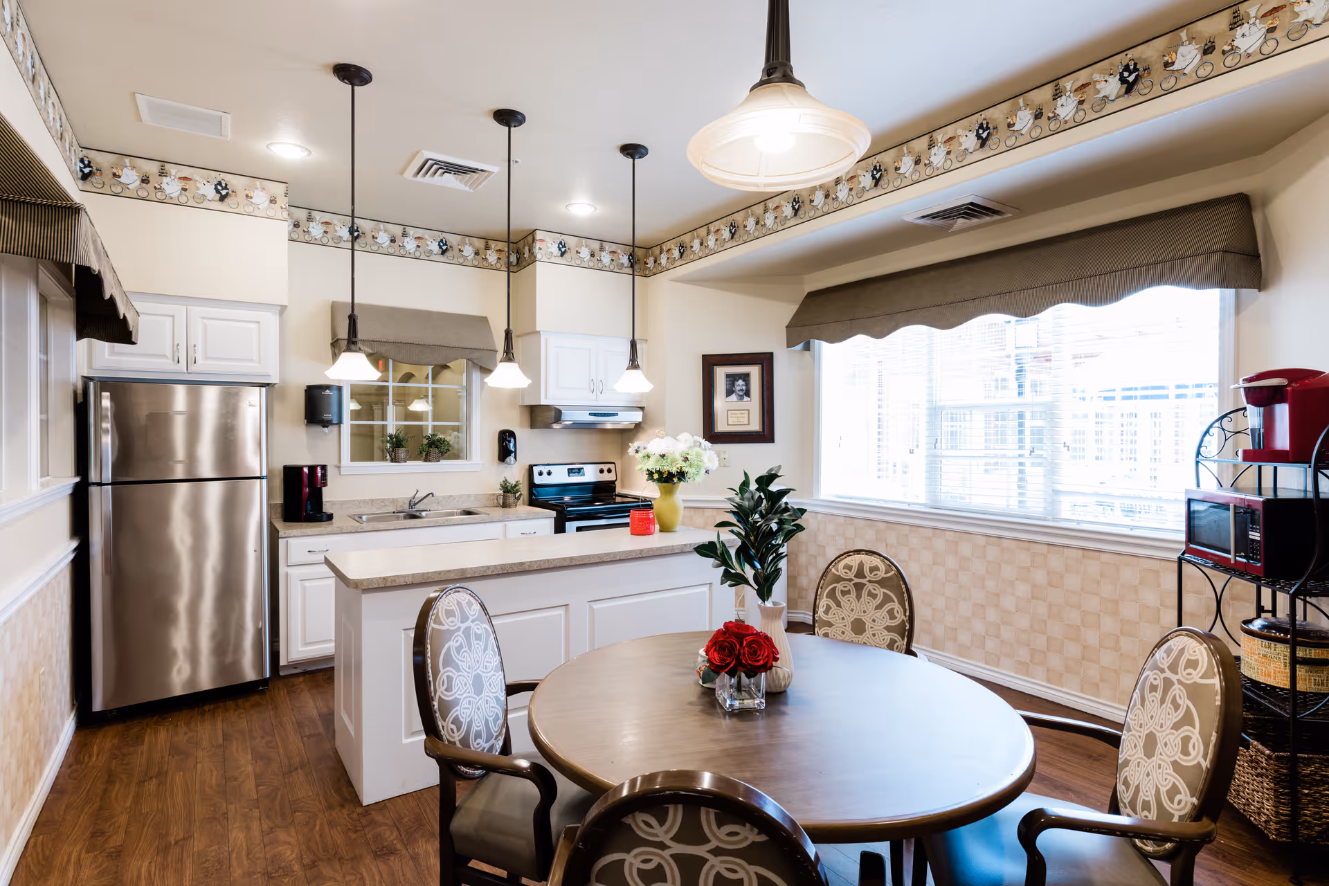 A bright kitchen and dining area with a round wooden table surrounded by four patterned chairs. The kitchen features white cabinets, a stainless steel refrigerator, an electric stove, and a countertop island with a vase of flowers. Three pendant lights hang above the island. A large window with blinds lets in natural light, and a coffee maker and microwave are visible on a black metal rack.