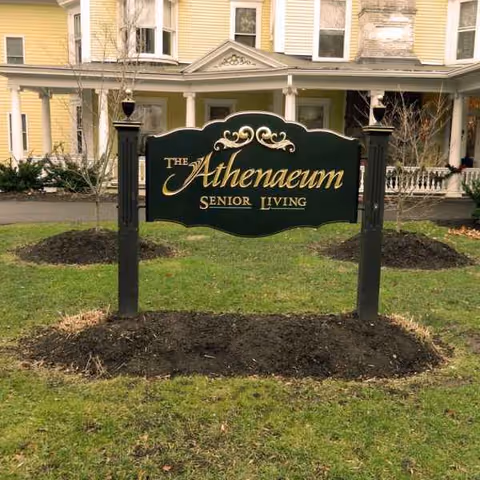 A decorative sign reading The Athenaeum Senior Living stands on a grassy front yard.