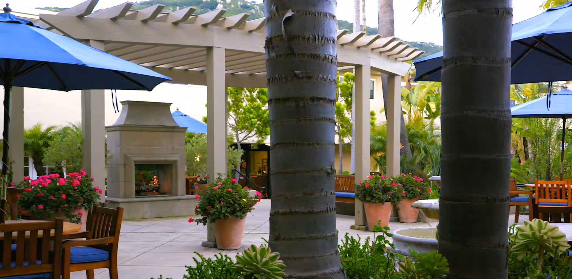 Outdoor courtyard with a pergola and fireplace, palm trees, patio tables with blue umbrellas, and potted flowering plants.