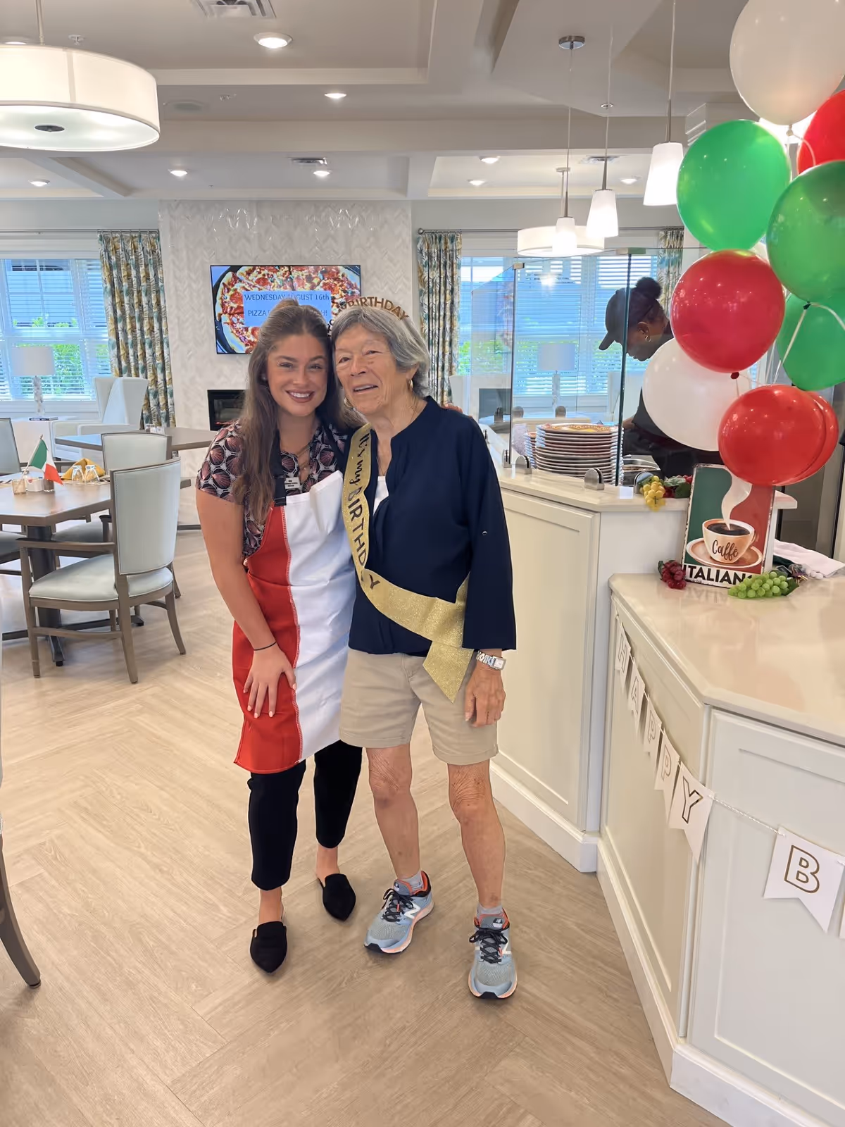 A young woman wearing a red and white apron poses and smiles next to an elderly woman wearing a 'Happy Birthday' sash and headband inside a bright dining area decorated with balloons and a 'Happy Birthday' banner. In the background, a person is preparing food behind a counter with plates stacked, and a TV screen displays a pizza event announcement.