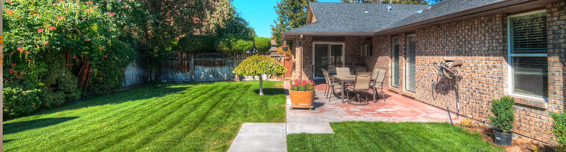 A well-maintained backyard with a green lawn, a small tree with a rounded top, and a patio area attached to a brick building. The patio has a table with six chairs and a large potted plant. The area is surrounded by a wooden fence and various bushes and flowering plants. The sky is clear and blue.