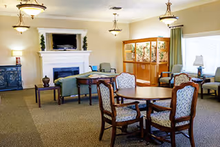 A spacious and well-lit living room area with a fireplace, a TV mounted above it, several upholstered chairs and sofas, a wooden table with four cushioned chairs, and large windows with curtains allowing natural light to enter.