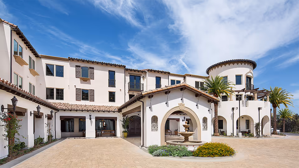 Exterior view of Crestavilla senior living facility featuring a Mediterranean-style building with white stucco walls, terracotta roof tiles, arched doorways, a central fountain, palm trees, and a clear blue sky with some clouds.
