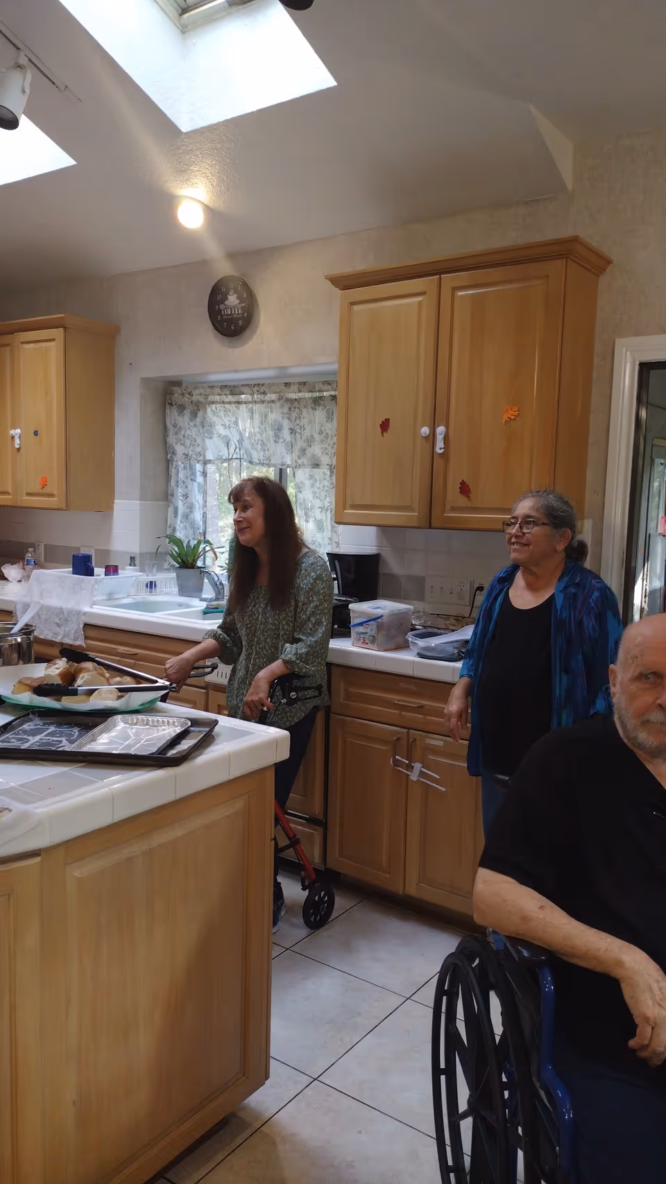 A kitchen scene with three elderly people. One woman is standing near the kitchen counter holding a spatula, another woman stands nearby smiling, and a man in a wheelchair is partially visible in the foreground. The kitchen has wooden cabinets, a white tiled countertop, and a window with floral curtains. There are skylights on the ceiling providing natural light.