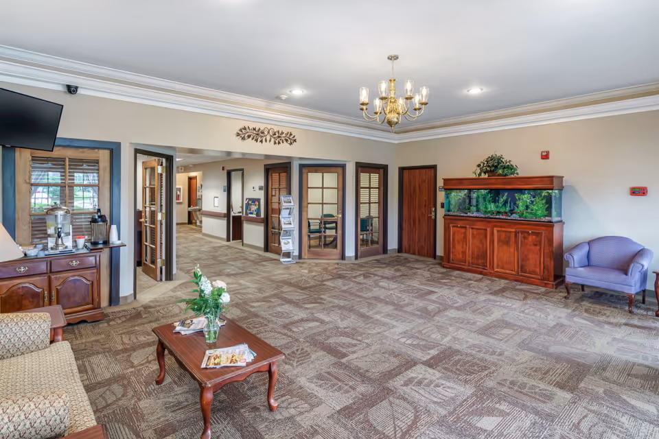 A spacious and well-lit common area in a senior living facility featuring a patterned carpet, a wooden coffee table with a flower vase and magazines, a sofa, a purple armchair, a large aquarium on a wooden stand, and a chandelier hanging from the ceiling. There are multiple doors and windows with wooden frames, a water dispenser on a wooden cabinet, and a hallway leading to other rooms.