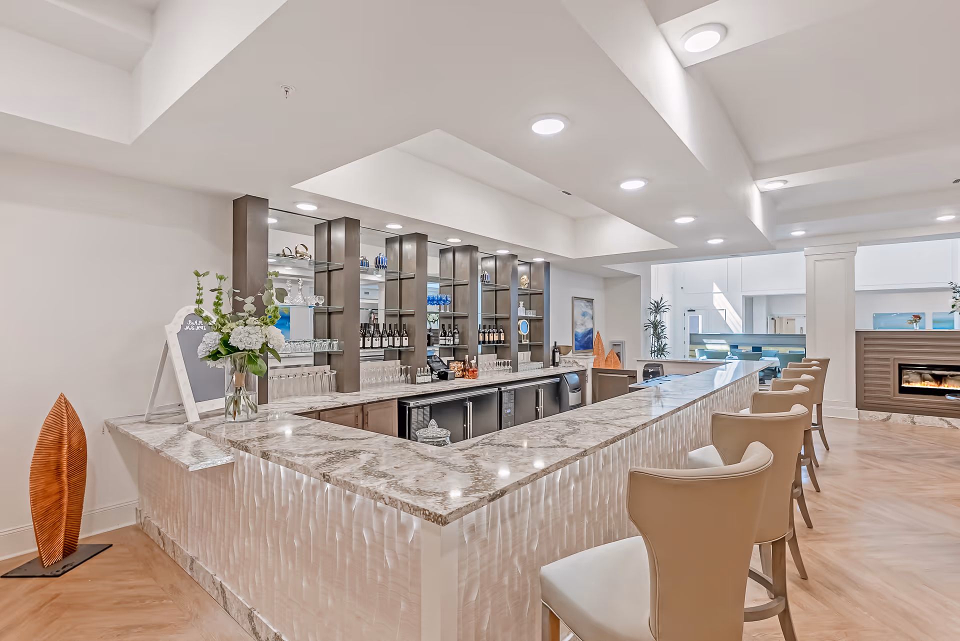 A modern bar area with a marble countertop and several beige bar stools. Behind the bar are shelves stocked with wine bottles, glasses, and decorative items. The space is well-lit with recessed ceiling lights and features light-colored walls and wood flooring. There is a fireplace visible in the background and some plants and artwork decorating the area.