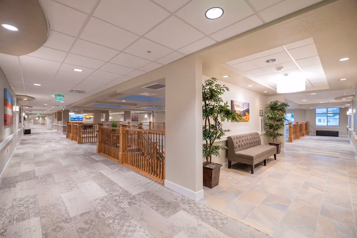 A spacious and well-lit hallway in a senior living facility with light-colored walls and carpeted floors. There is a seating area with a cushioned bench and potted plants near a tiled floor section. Wooden railings surround an open area in the middle, and framed artwork decorates the walls. Ceiling lights provide bright illumination throughout the corridor.