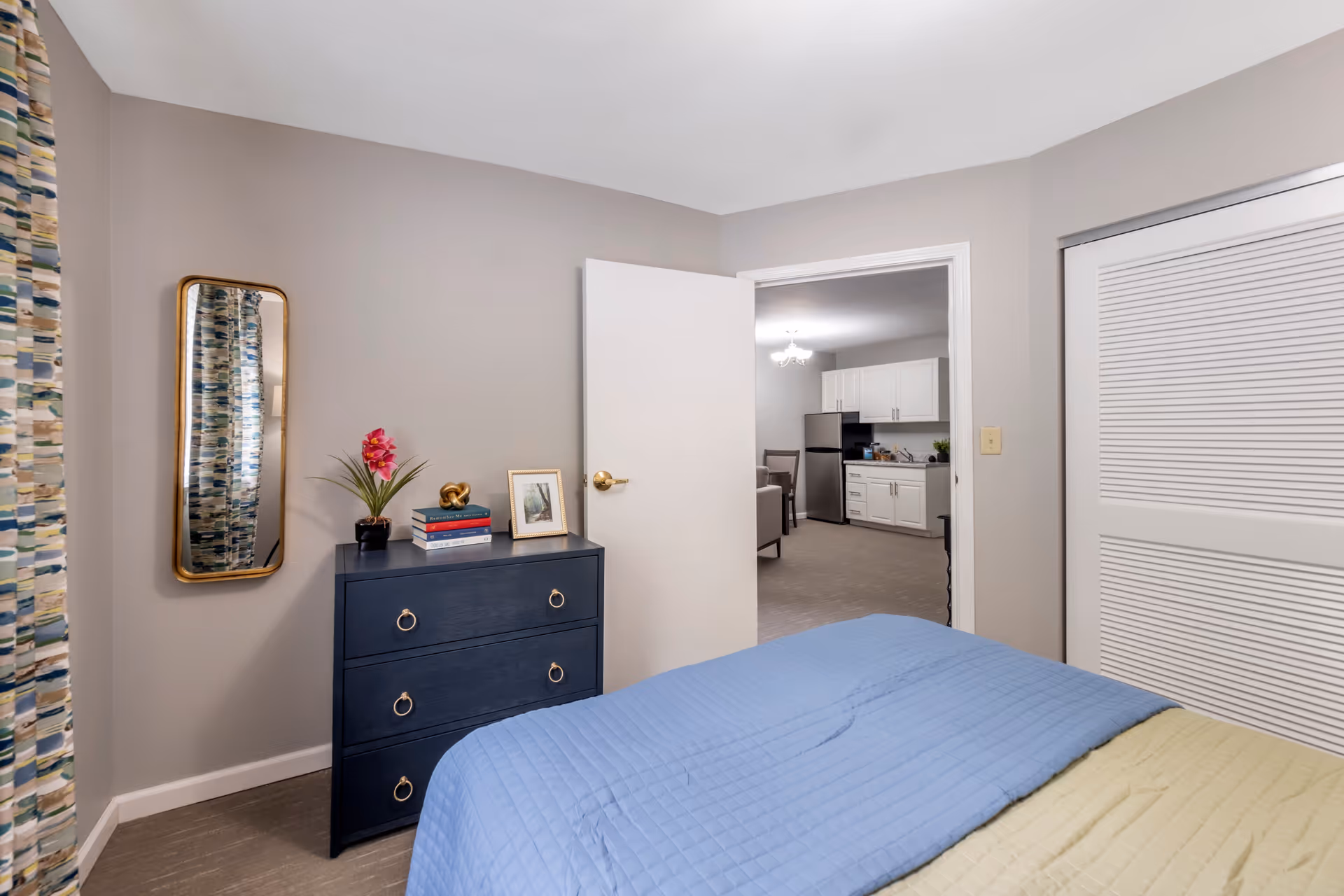 A furnished bedroom with a blue quilted bed, navy dresser topped with decor, a wall mirror, and an open door looking into a kitchen/living area.