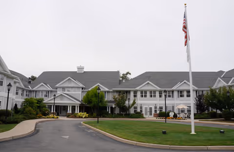 Exterior view of a large senior living facility building with white siding and gray roofs, surrounded by landscaped greenery and a circular driveway with an American flag on a flagpole in the center.