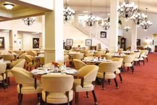 A spacious dining room with multiple round tables set with white tablecloths, plates, and utensils. Each table is surrounded by beige upholstered chairs. The room features red carpeting, white walls with columns, and elegant chandeliers hanging from the ceiling. Artwork is displayed on the walls.