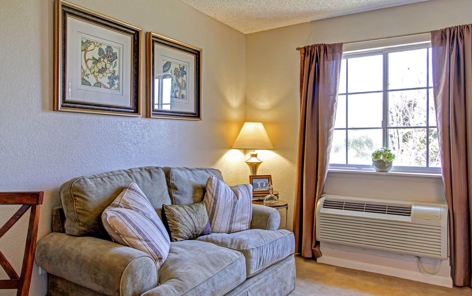A cozy living room area with a gray cushioned loveseat adorned with three pillows. Behind the loveseat, two framed floral artworks hang on the wall. To the right, a table lamp on a small round side table provides warm light. A window with brown curtains lets in natural light, and below it is a wall-mounted air conditioning unit.