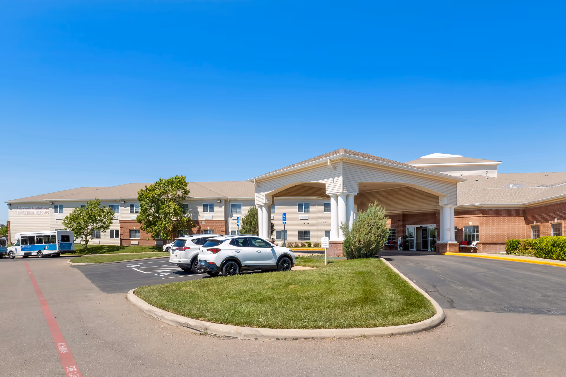 Exterior view of Brookdale Medi Park West senior living facility showing the main entrance with a covered drop-off area, several parked cars, a shuttle bus, and a clear blue sky.