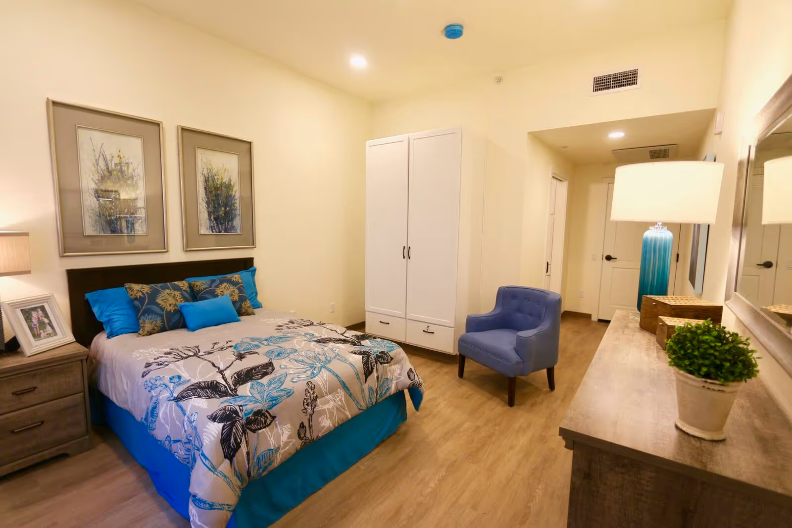 A well-lit bedroom with a bed featuring blue and floral bedding, two framed floral artworks above the headboard, a wooden nightstand with a lamp and a framed photo, a white wardrobe, a blue upholstered chair, a wooden dresser with a lamp, decorative boxes, and a small potted plant, and a large mirror on the wall.