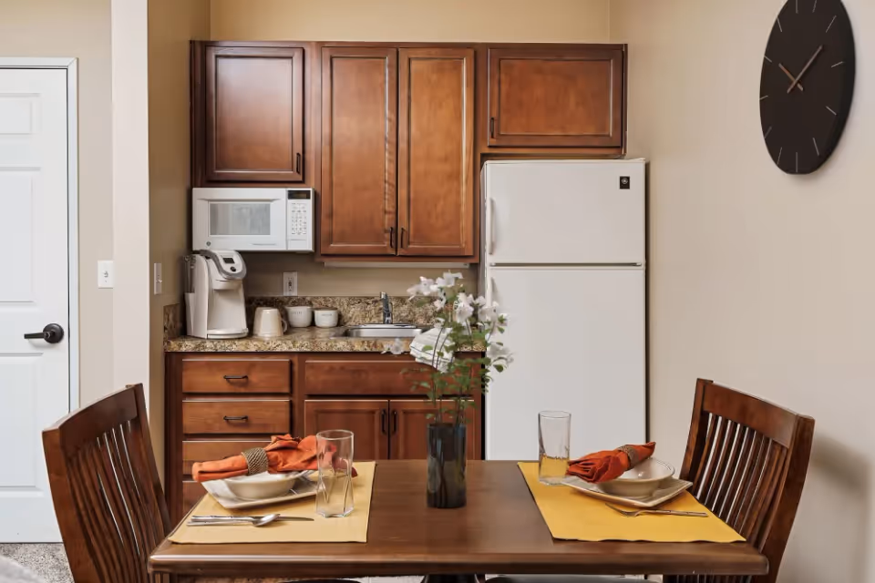 Small kitchen with wooden cabinets, a white refrigerator and a dining table set for two with place settings and a vase of flowers.