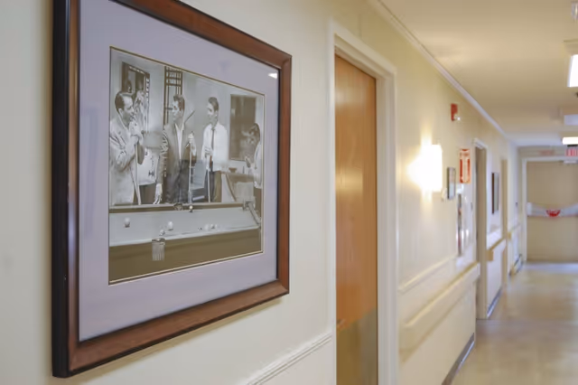 A hallway in a senior living facility with beige walls, handrails, and several closed wooden doors. A framed black and white photograph of four men playing pool is hung on the left wall. The hallway is well-lit with wall-mounted lights and ceiling fixtures.
