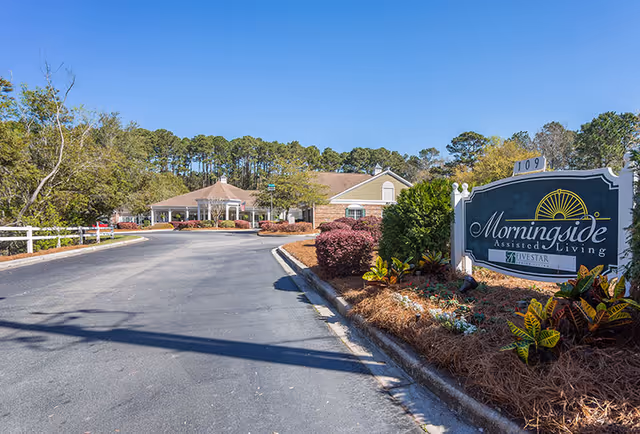Entrance driveway leading to a single-story assisted living facility building surrounded by trees and landscaped bushes. A large sign on the right side reads 'Morningside Assisted Living' with the number 109 on top.