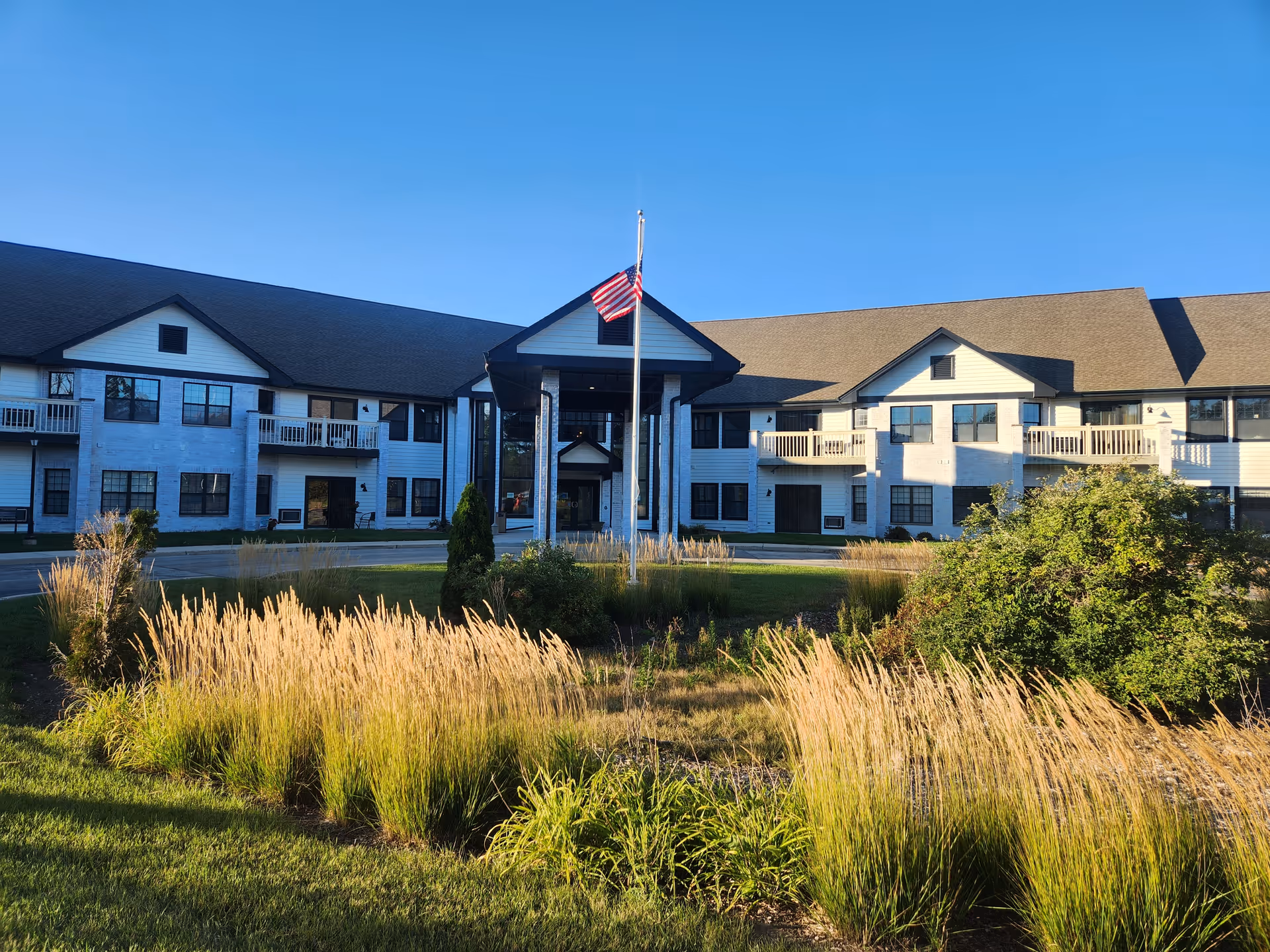 Exterior front view of a two-story senior living building with an American flag at the entrance and landscaped ornamental grasses.