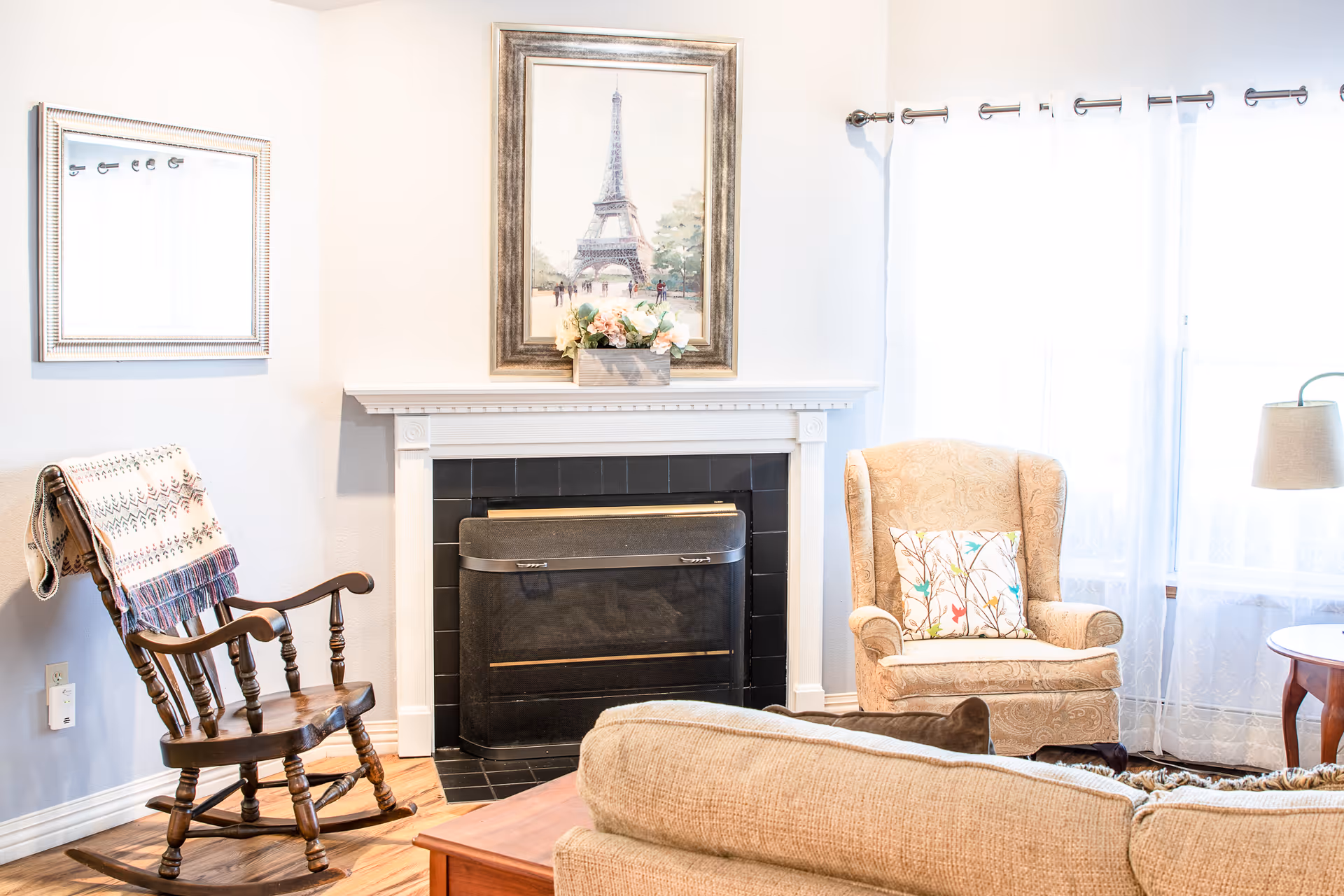 Bright living room with a fireplace topped by a framed Eiffel Tower print, a patterned armchair, wooden rocking chair, and a sofa.