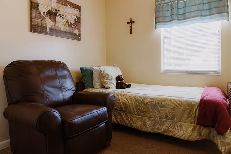 A small bedroom with a single bed covered in a light-colored patterned bedspread, a red blanket folded at the foot, and a teddy bear sitting on the pillows. Next to the bed is a large brown leather armchair. A window with blinds and a blue valance lets in natural light. On the wall above the bed is a wooden cross and a framed world map artwork.