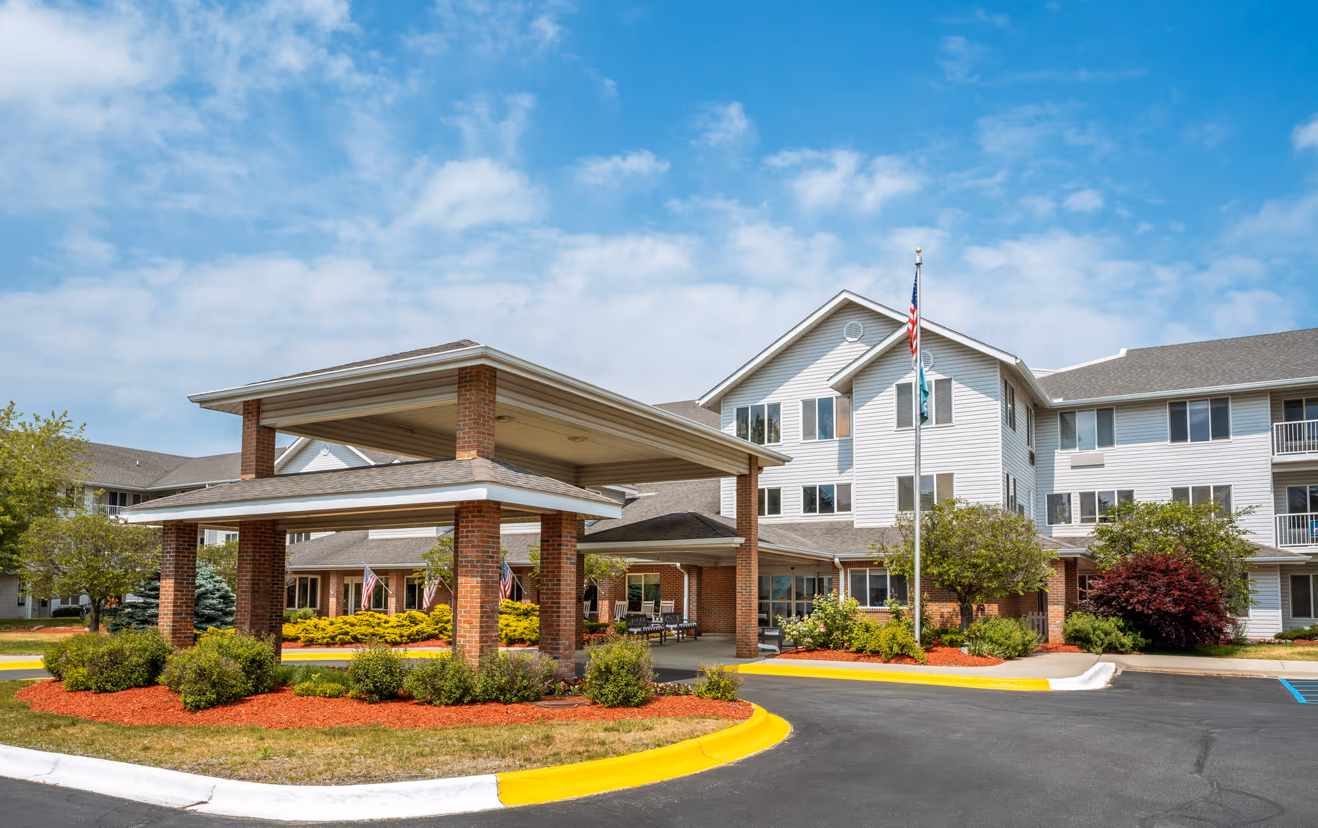 Exterior view of Holiday Blue Water Lodge, a multi-story senior living facility with white siding and brick accents. The entrance features a covered drop-off area supported by brick columns. The surrounding landscaping includes bushes, small trees, and mulch beds. An American flag and another flag are displayed on a flagpole near the entrance. The sky is partly cloudy.