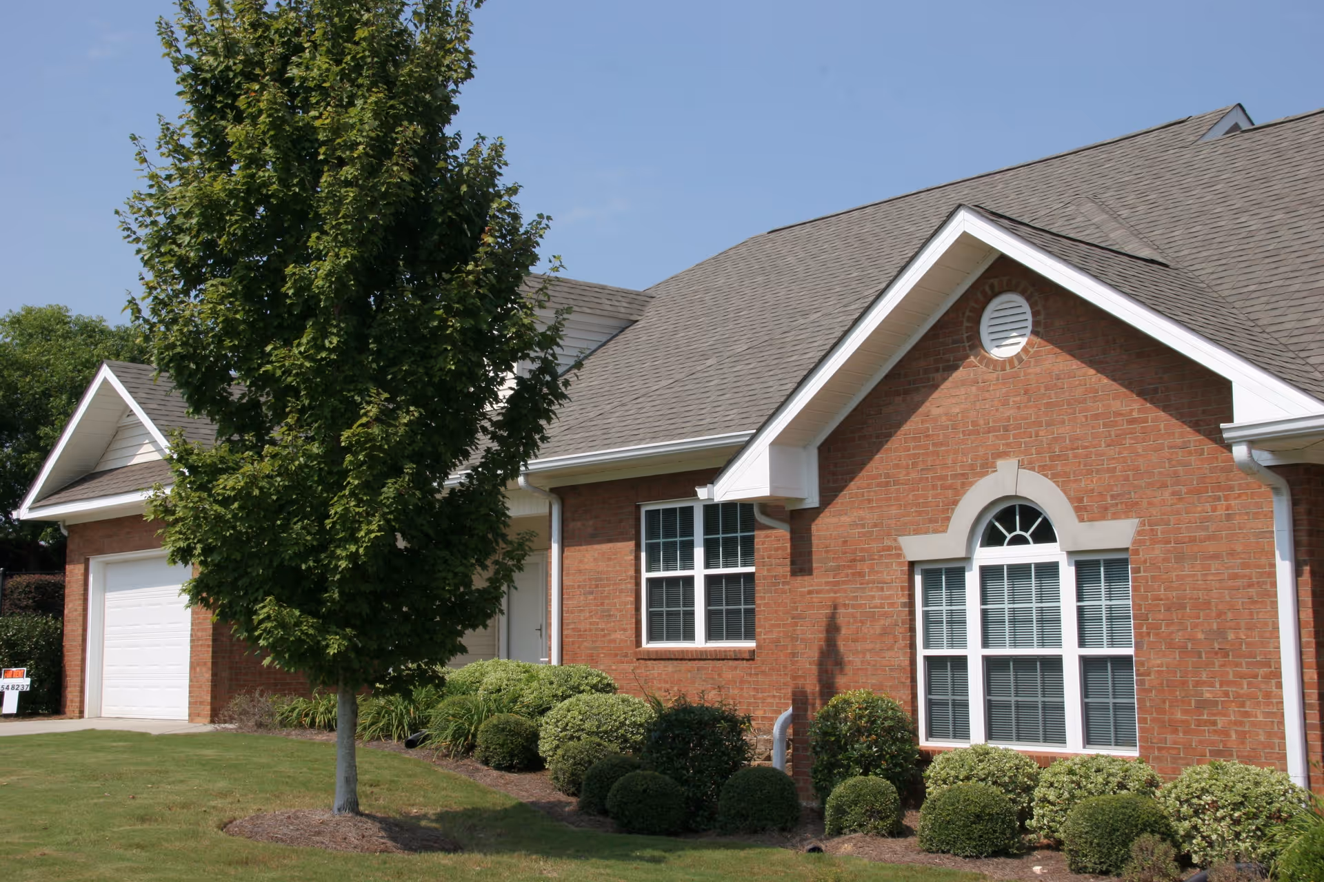 Exterior view of a single-story brick building with a gray shingled roof, white-trimmed windows, and a garage door. There is a tree and neatly trimmed bushes in the front yard under a clear blue sky.